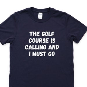 Może przedstawiać: Koszulka w kolorze granatowym z białym napisem "The golf course is calling and I must go."