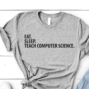 May include: A grey t-shirt with the text "Eat. Sleep. Teach Computer Science." printed on it. The shirt is laid out on a white wooden surface with a pair of white trainers and a pair of blue jeans.