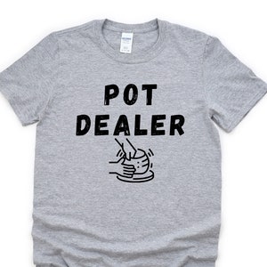 May include: A grey t-shirt with the text "POT DEALER" in black lettering. The text is accompanied by a black and white illustration of hands shaping clay on a pottery wheel.