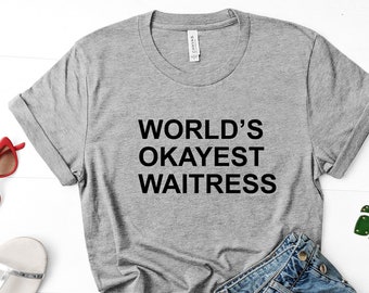 May include: A gray t-shirt with the text "WORLD'S OKAYEST WAITRESS" is displayed with denim shorts, red sunglasses, silver sandals, and a green leaf keychain. The shirt is knotted at the waist, suggesting a casual style.