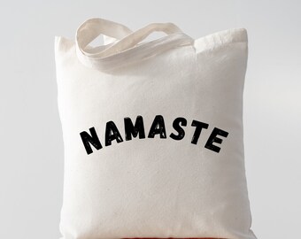 May include: A white canvas tote bag with black text that reads "NAMASTE" in a bold, stylized font. The bag is sitting on a wooden stool.