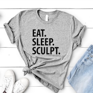 May include: A heather gray t-shirt with the words "EAT. SLEEP. SCULPT." in black block letters. The shirt is styled with light blue jeans and white sneakers. Small potted succulents are arranged on a white wooden surface.