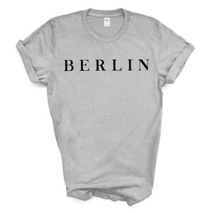May include: A light gray t-shirt with the word "BERLIN" printed in black, centered on the chest. The sleeves are rolled up, and the shirt is displayed on a white background. The t-shirt is a crew neck style.