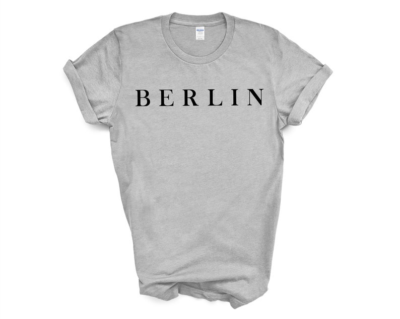 May include: A light gray t-shirt with the word "BERLIN" printed in black, centered on the chest. The sleeves are rolled up, and the shirt is displayed on a white background. The t-shirt is a crew neck style.