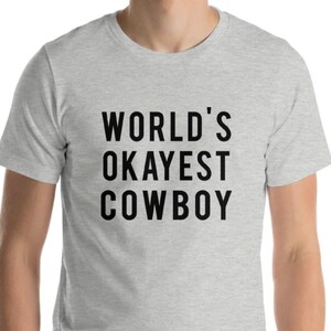 May include: A light gray t-shirt with the text "WORLD'S OKAYEST COWBOY" in bold black letters. The shirt is made of a soft material and has short sleeves.