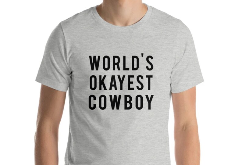 May include: A light gray t-shirt with the text "WORLD'S OKAYEST COWBOY" in bold black letters. The shirt is made of a soft material and has short sleeves.