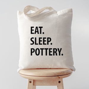 May include: A white canvas tote bag with black text that reads "EAT. SLEEP. POTTERY." The bag is sitting on a wooden stool.