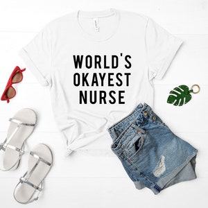 May include: White t-shirt with black text that reads "WORLD'S OKAYEST NURSE". The shirt is laid flat on a white wooden surface with a pair of denim shorts, sunglasses, and a green leaf keychain.