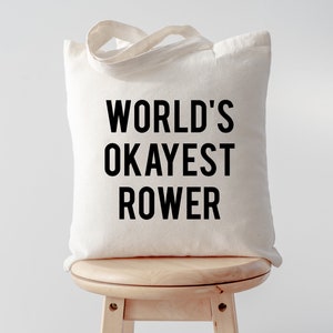 Puede incluir: Una bolsa de lona blanca con texto negro que dice "WORLD'S OKAYEST ROWER". La bolsa está sentada en un taburete de madera.