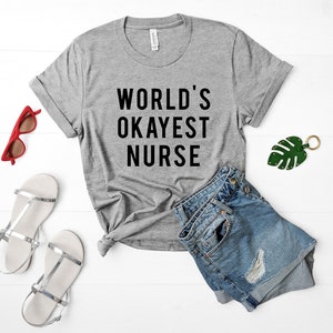 May include: A gray t-shirt with the text "WORLD'S OKAYEST NURSE" printed in black. The shirt is laid flat on a white surface with a pair of denim shorts, sunglasses, and a green leaf keychain.