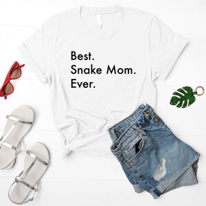 May include: White t-shirt with the text "Best. Snake Mom. Ever." on a white background. The shirt is laying on a white surface with a pair of denim shorts, a pair of silver sandals, and a green leaf keychain.