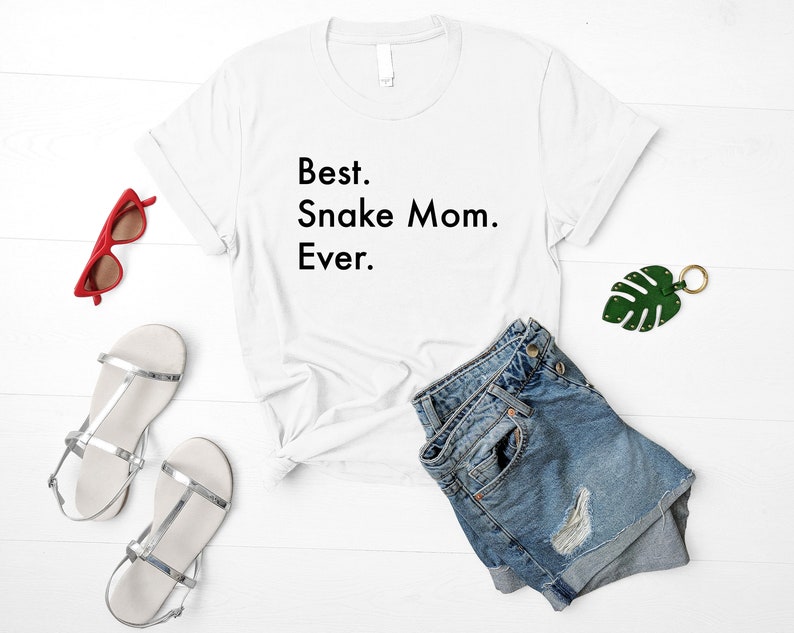 May include: White t-shirt with the text "Best. Snake Mom. Ever." on a white background. The shirt is laying on a white surface with a pair of denim shorts, a pair of silver sandals, and a green leaf keychain.