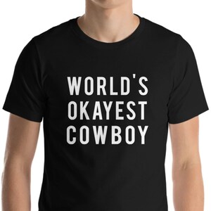 May include: Black t-shirt with the white text "WORLD'S OKAYEST COWBOY". The shirt is made of a soft material and has short sleeves. The text is centered on the front of the shirt.