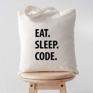 Op de afbeelding: Een crèmekleurige draagtas met de woorden "EAT. SLEEP. CODE." gedrukt in vette zwarte letters. De tas heeft een eenvoudig ontwerp met twee handvatten en staat op een houten kruk.