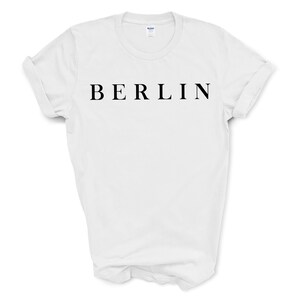 May include: White t-shirt with the word "BERLIN" printed in black, centered across the chest. The shirt has short sleeves and a crew neck. The sleeves are rolled up. The background is plain white.