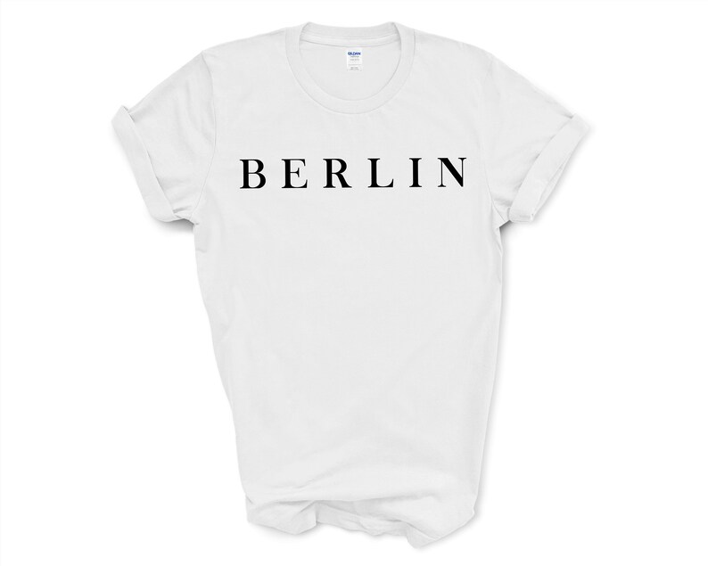 May include: White t-shirt with the word "BERLIN" printed in black, centered across the chest. The shirt has short sleeves and a crew neck. The sleeves are rolled up. The background is plain white.
