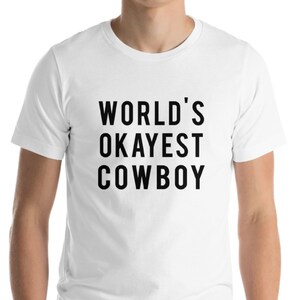May include: White t-shirt with the text "WORLD'S OKAYEST COWBOY" in black, bold letters. The shirt is a short-sleeved crew neck style.