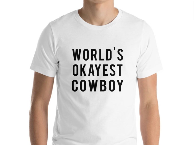 May include: White t-shirt with the text "WORLD'S OKAYEST COWBOY" in black, bold letters. The shirt is a short-sleeved crew neck style.