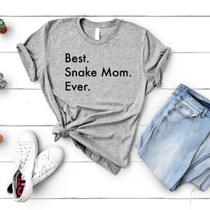 May include: A gray t-shirt with the text "Best. Snake Mom. Ever." printed on it. The shirt is laid out on a white wooden surface with a pair of white sneakers, a pair of blue jeans, and a few small potted plants.