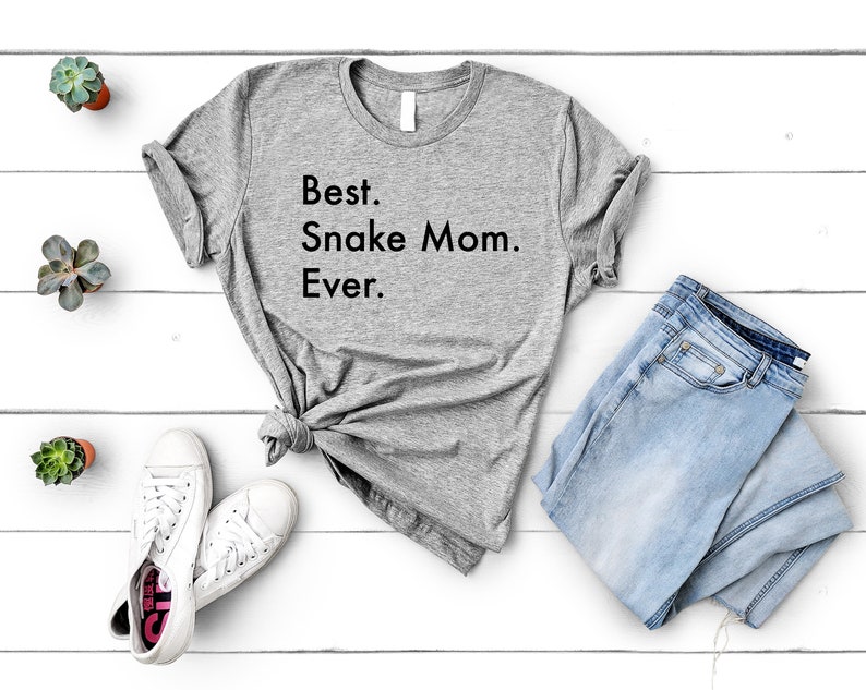 May include: A gray t-shirt with the text "Best. Snake Mom. Ever." printed on it. The shirt is laid out on a white wooden surface with a pair of white sneakers, a pair of blue jeans, and a few small potted plants.