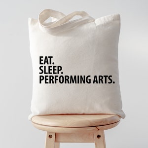 Puede incluir: Una bolsa de tela blanca con texto negro que dice "EAT. SLEEP. PERFORMING ARTS."