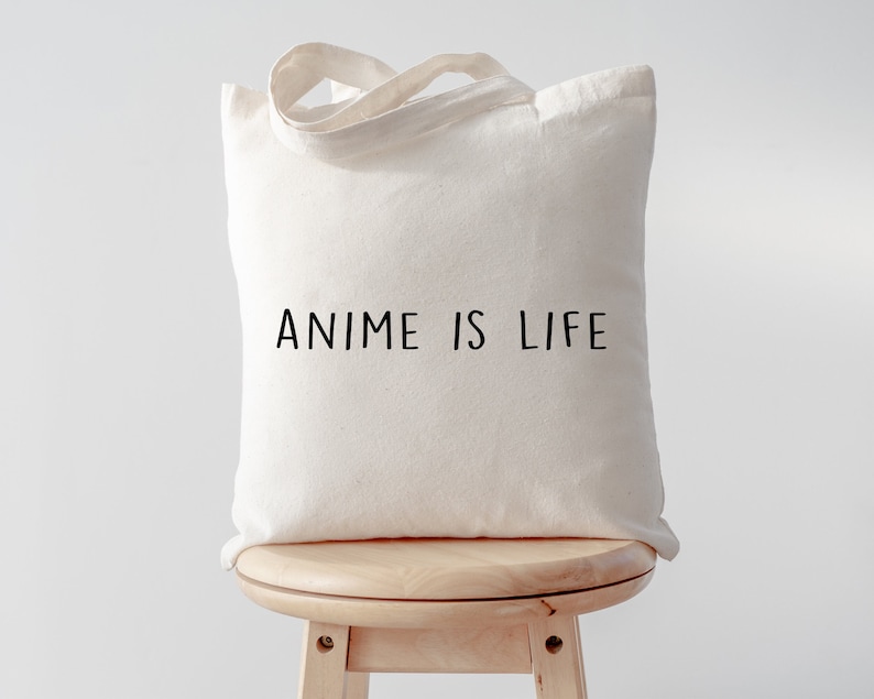 May include: A white canvas tote bag with black text that reads "Anime is Life". The bag is sitting on a wooden stool.