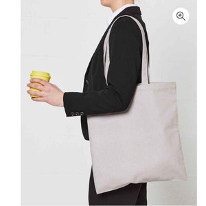 May include: A person holding a yellow reusable coffee cup and carrying a large, light grey tote bag with long straps.