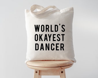 以下が含まれることがあります： 白いキャンバス地のトートバッグに、黒い文字で「WORLD'S OKAYEST DANCER」と書いてあります。バッグは木製の踏み台の上に置かれています。