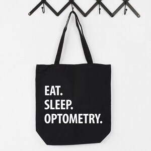 May include: Black canvas tote bag with white text that reads "Eat. Sleep. Optometry."
