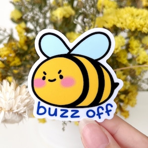 Könnte beinhalten: Ein weißer Aufkleber mit einer Cartoon-Biene mit schwarzen Streifen und blauen Flügeln. Die Biene hat einen gelben Körper und einen mürrischen Gesichtsausdruck. Der Text "buzz off" ist in Blau unter der Biene geschrieben.