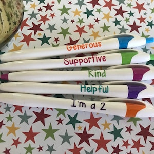May include: Six white pens with colorful accents and positive messages written on them. The messages are "Generous", "Supportive", "Kind", "Helpful", and "I'm a 2". The pens are arranged in a stack on a background of colorful stars.