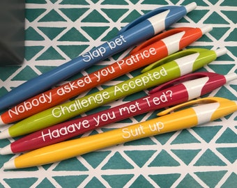 May include: Five colorful pens with white caps and white text on a teal and white patterned background. The text on the pens reads: "Slap bet", "Nobody asked you Patrice", "Challenge Accepted", "Haaave you met Ted?", and "Suit up".