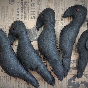 May include: Five black felt crow ornaments with red bead eyes. The crows are arranged in a row on a patterned fabric background.