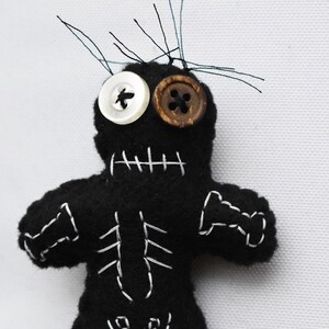 May include: Black felt voodoo doll with white stitching, button eyes, and a stitched-on smile. The doll has a skeleton design on its front and is standing on a white surface.