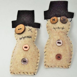 May include: Two felt snowman ornaments with black top hats and button eyes. One snowman has a brown button nose and the other has a pink button nose.