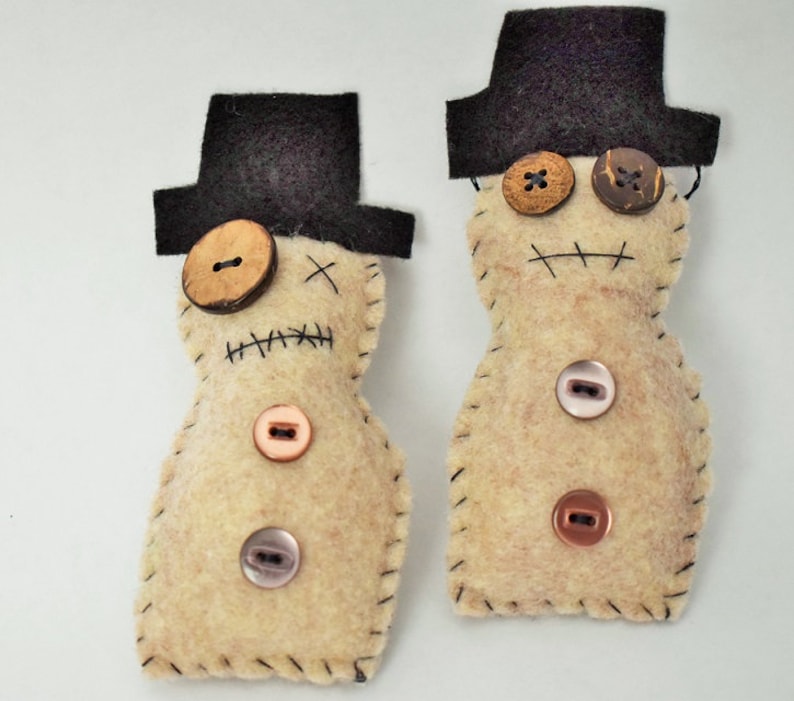 May include: Two felt snowman ornaments with black top hats and button eyes. One snowman has a brown button nose and the other has a pink button nose.