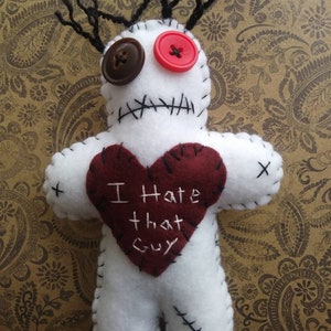 May include: A handmade white voodoo doll with black stitched details and yarn hair. It has a brown button and a red button for eyes. A burgundy heart is sewn on the front with the text "I Hate that Guy".