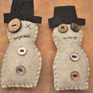 May include: Two felt doll ornaments with black top hats and stitched details. The dolls have brown button eyes and a stitched mouth. One doll has a brown button nose and the other has a wooden button nose. The dolls are decorated with various buttons.
