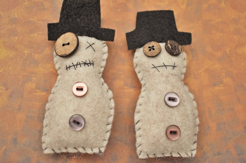 May include: Two felt doll ornaments with black top hats and stitched details. The dolls have brown button eyes and a stitched mouth. One doll has a brown button nose and the other has a wooden button nose. The dolls are decorated with various buttons.