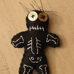 May include: Black felt voodoo doll with white stitching, a white button for an eye, and a brown button for the other eye.