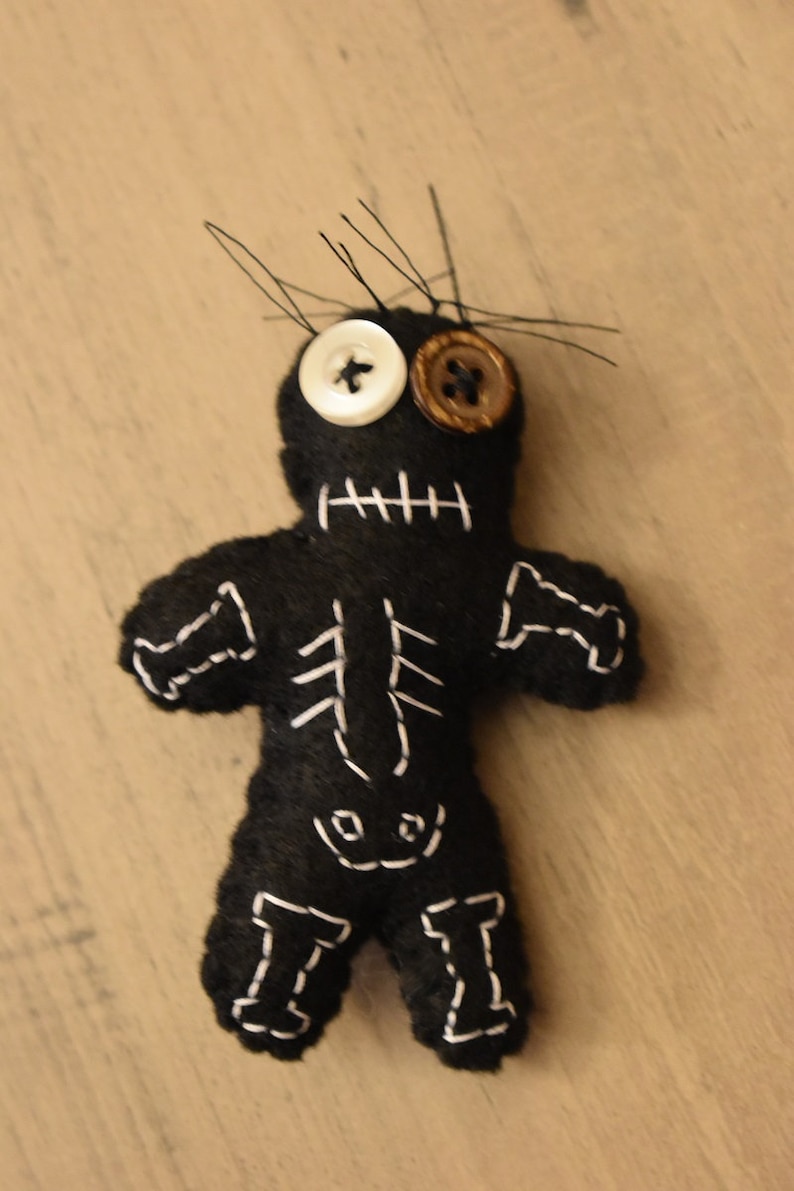 May include: Black felt voodoo doll with white stitching, a white button for an eye, and a brown button for the other eye.