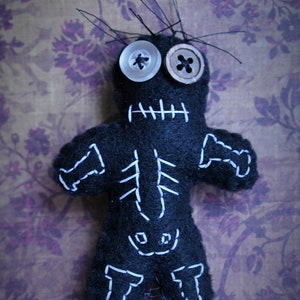 May include: A black voodoo doll with white stitching outlining a skeleton, with two buttons for eyes. The doll is on a purple floral background.