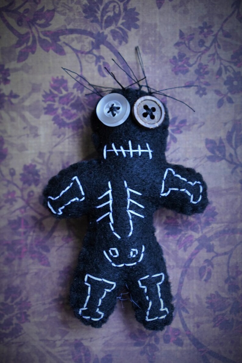 May include: A black voodoo doll with white stitching outlining a skeleton, with two buttons for eyes. The doll is on a purple floral background.