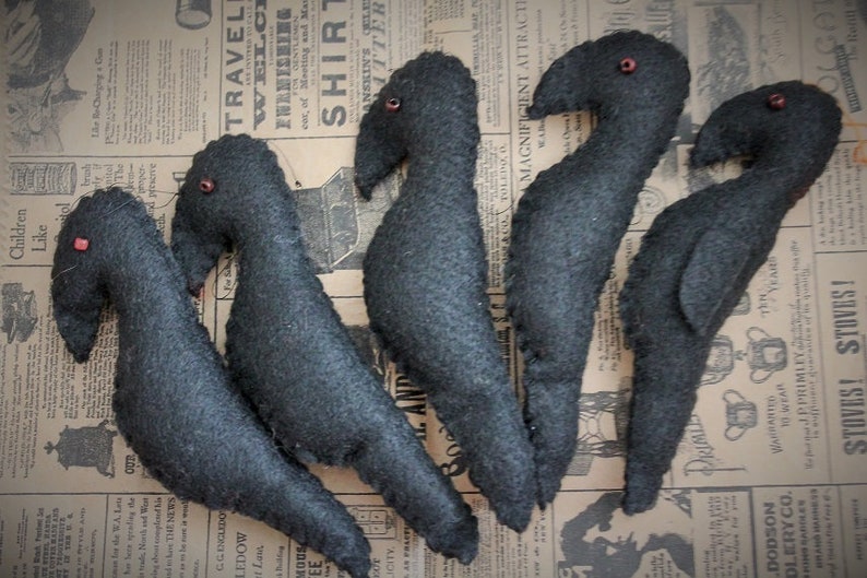 May include: Five black felt crow ornaments with red bead eyes. The crows are arranged in a row on a patterned fabric background.