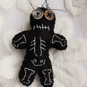 May include: A black felt voodoo doll with white stitching, button eyes, and a stitched-on mouth. The doll is lying on a white lace surface.
