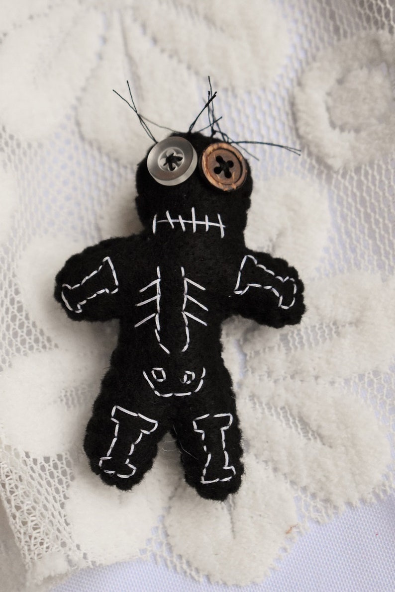 May include: A black felt voodoo doll with white stitching, button eyes, and a stitched-on mouth. The doll is lying on a white lace surface.