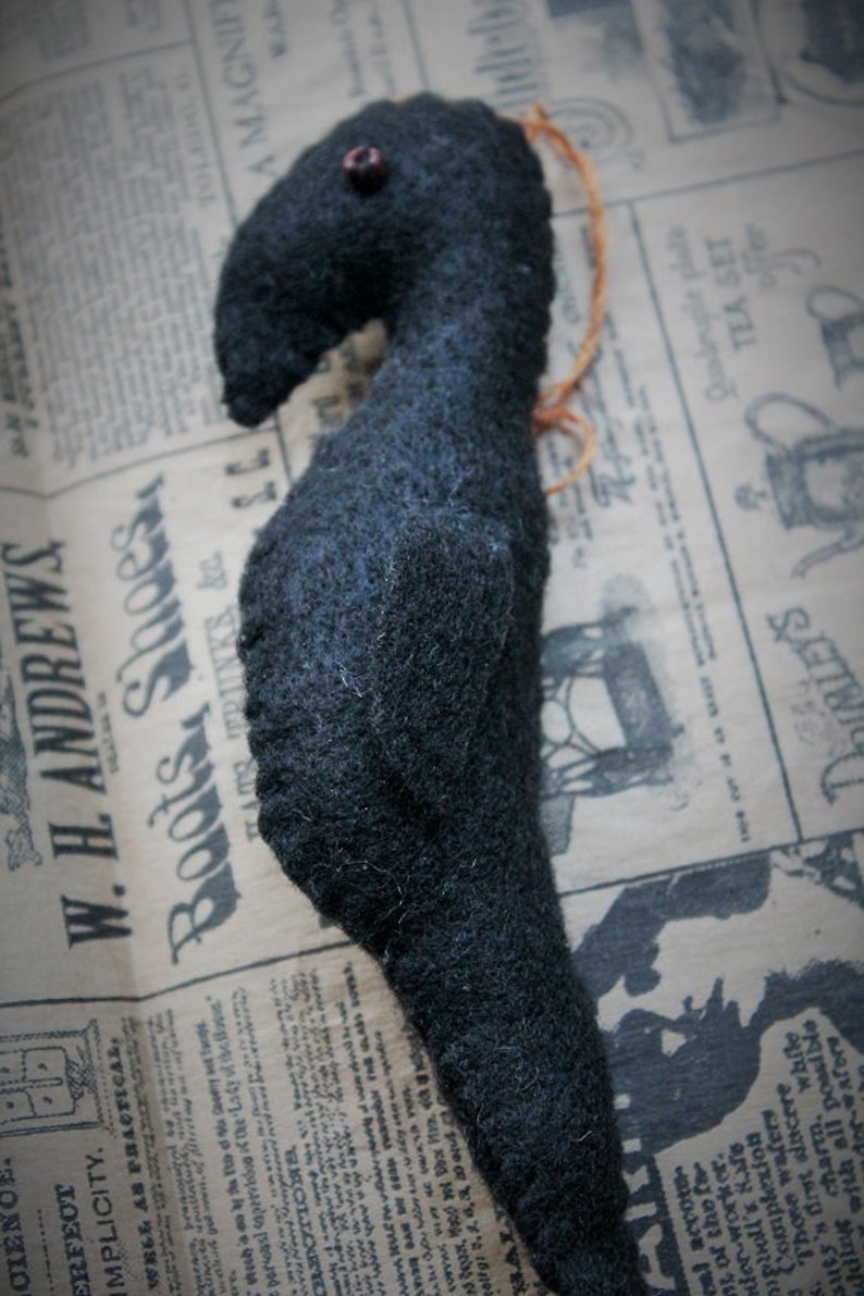 May include: A black felt crow with a red bead for an eye. The crow is sitting on a piece of old newspaper.