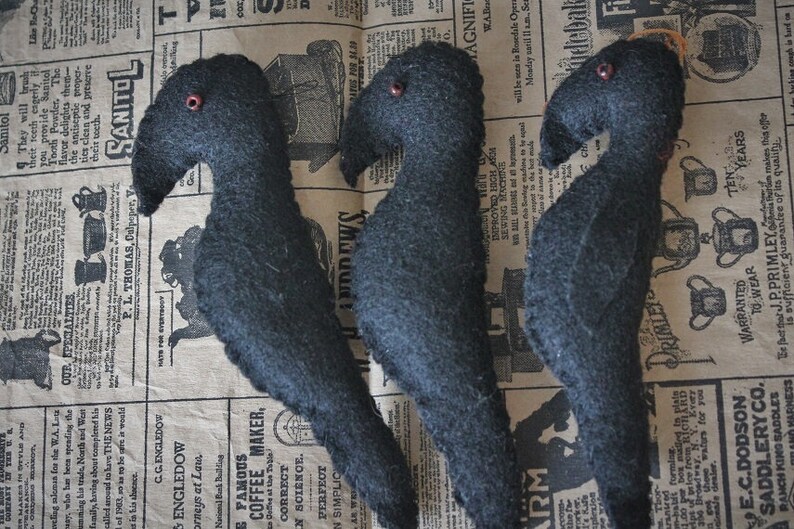 May include: Three black felt crow figurines with red bead eyes. The crows are arranged in a row on a vintage newspaper background.