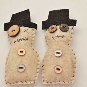 May include: Two felt dolls with black hats and button eyes. The dolls have stitched mouths and are decorated with various buttons.