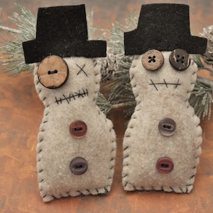 May include: Two felt snowman ornaments with black top hats, stitched details, and various button eyes and accents.